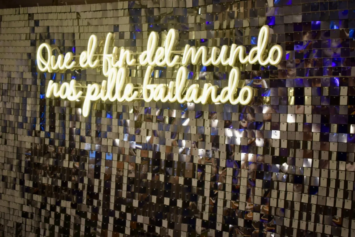 Neón LED «Que el fin del mundo nos pille bailando» sobre la pared de lentejuelas